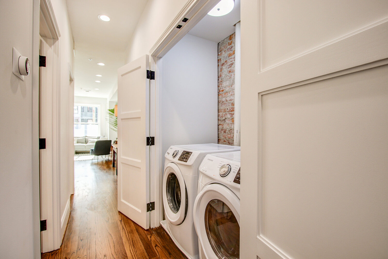 Hallway in San Diego home featuring a laundry closet with front-load washer and dryer, exposed brick wall, and wood flooring leading to a bright sitting area
