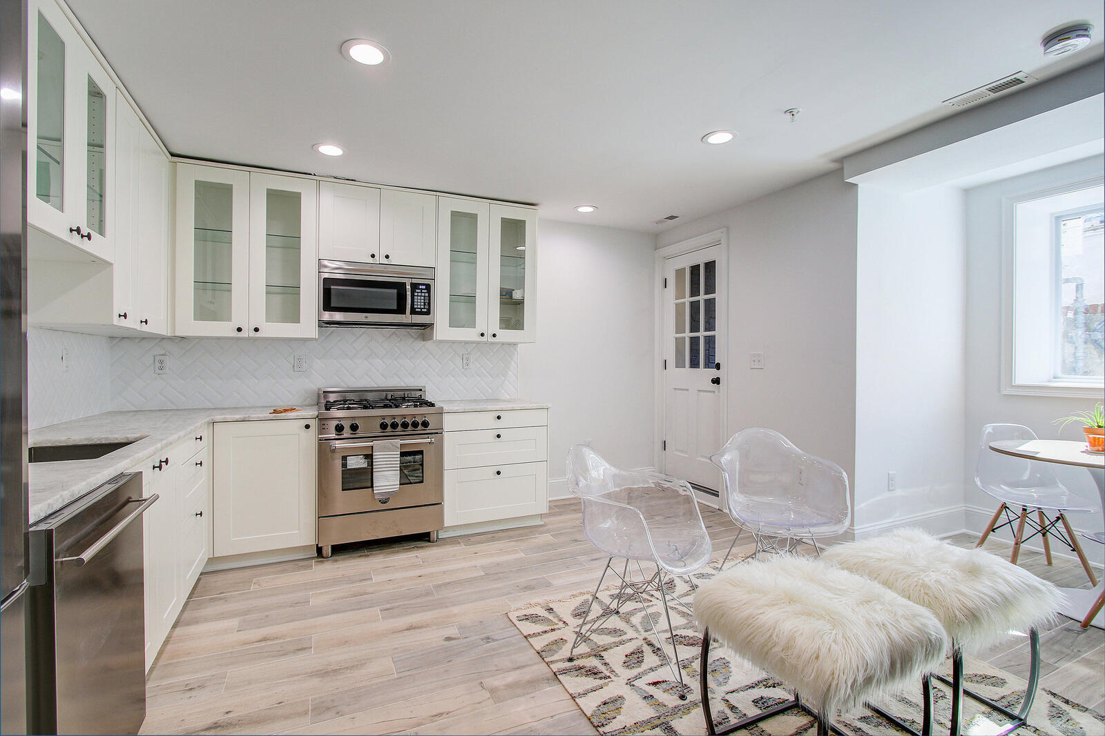 Modern white kitchen in San Diego featuring glass cabinet doors, stainless steel appliances, and cozy seating with natural light